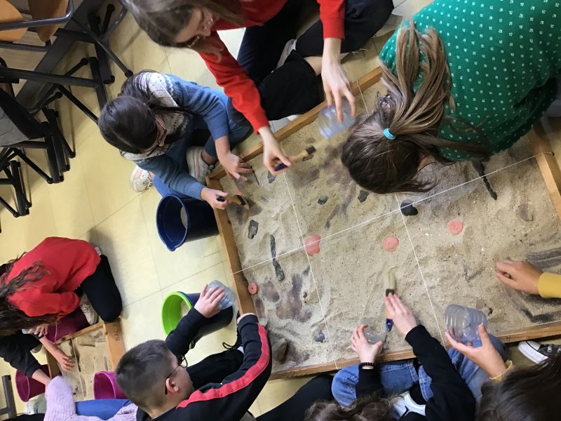 Des élèves dégagent des objets dans un bac de fouilles quadrillé lors d’un atelier Archéoskol.